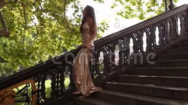 迷人的年轻健康的女人穿着一件金色闪亮的长裙子，在夏天的高跟鞋上走下楼梯。 缔约方